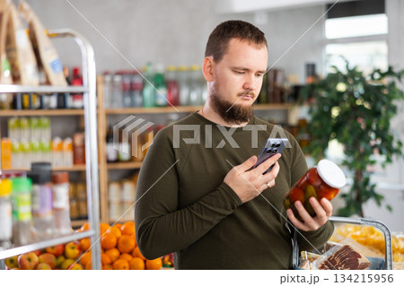 Male shopper scans a QR code on a label of canned cucumbers and tomatos in supermarket 134215956