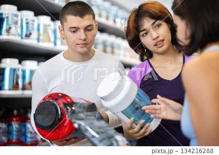 Young guy and women choosing protein powder jar in sports nutrition store Young guy and women choosing protein powder jar in sports nutrition store 134216288