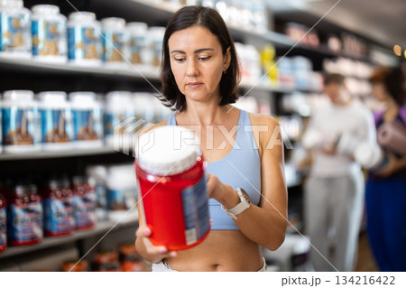 Focused woman choosing protein powder jar in sports nutrition store 134216422
