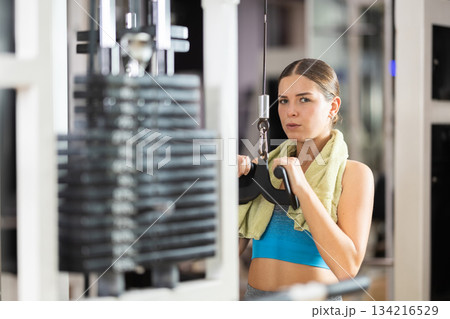 Girl in gym performs exercises for arm muscles using cable crossover machine and pull weighted rope Girl in gym performs exercises for arm muscles using cable crossover machine and pull weighted rope 134216529