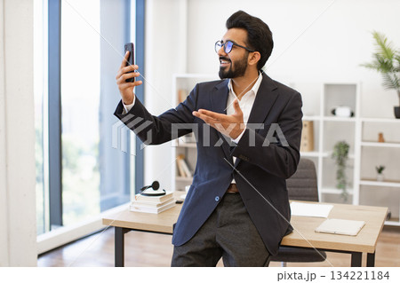 An Indian businessman smiles while on a video call in his office, gesturing with his hand An Indian businessman smiles while on a video call in his office, gesturing with his hand 134221184