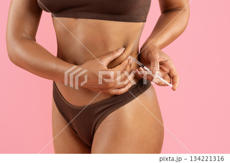Young black woman injecting syringe in belly fat area on pink background Young black woman injecting syringe in belly fat area on pink background 134221316