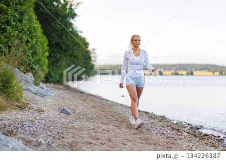 Young woman walking outdoors by a serene lake during a fitness routine 134226387