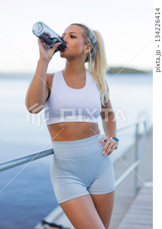 Active woman drinking water after outdoor workout beside a serene lake view 134226414