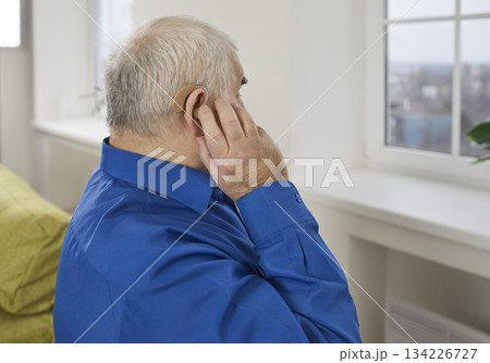 Side view of gray-haired senior man wearing modern hearing aid in his ear at home. 134226727