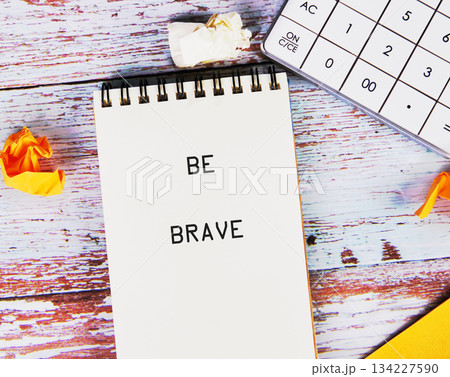 A notebook reads BE BRAVE, lying open on a wooden desk next to crumpled yellow and white paper. A calculator sits nearby, ready to help with bold financial decisions A notebook reads BE BRAVE, lying open on a wooden desk next to crumpled yellow and white paper. A calculator sits nearby, ready to help with bold financial decisions 134227590
