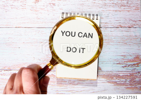 A hand holds a magnifying glass over a notebook. The text YOU CAN DO IT is shown enlarged through the glass. It is on a white washed wooden surface A hand holds a magnifying glass over a notebook. The text YOU CAN DO IT is shown enlarged through the glass. It is on a white washed wooden surface 134227591