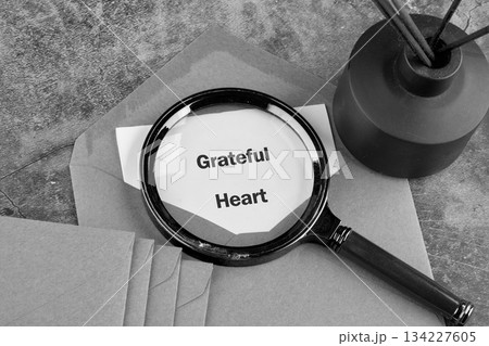 A magnifying glass rests on an envelope, focusing on the phrase Grateful Heart. A diffuser with reeds sits nearby on a textured stone surface A magnifying glass rests on an envelope, focusing on the phrase Grateful Heart. A diffuser with reeds sits nearby on a textured stone surface 134227605