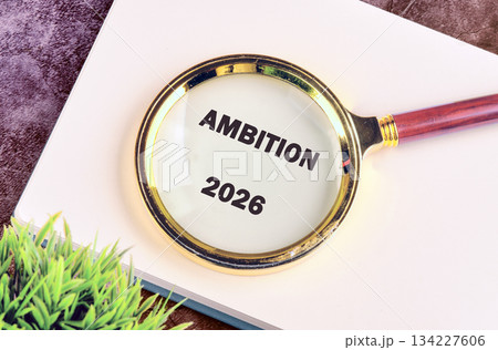 A magnifying glass focuses on the words AMBITION and 2026, sitting on a white book, next to a small green plant, suggesting careful planning and future goals 134227606