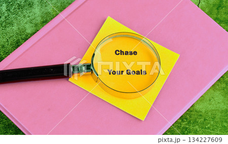 A magnifying glass highlights the phrase Chase Your Goals written on a vibrant yellow sticky note, set atop a cheerful pink book on a green surface A magnifying glass highlights the phrase Chase Your Goals written on a vibrant yellow sticky note, set atop a cheerful pink book on a green surface 134227609