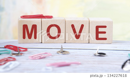 Wooden blocks with red lettering are arranged to spell MOVE. Colorful paper clips and silver thumbtacks surround the blocks on a white surface 134227618