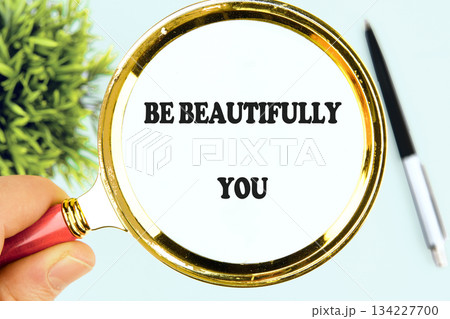 A hand holds a golden magnifying glass, focusing on the text BE BEAUTIFULLY YOU. A pen and verdant plant rest beside the sentiment, on a light blue surface A hand holds a golden magnifying glass, focusing on the text BE BEAUTIFULLY YOU. A pen and verdant plant rest beside the sentiment, on a light blue surface 134227700