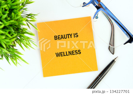 An inspiring note reads BEAUTY IS WELLNESS, sitting next to a lush green plant, sleek pen, and modern blue glasses, all resting on a pristine white surface An inspiring note reads BEAUTY IS WELLNESS, sitting next to a lush green plant, sleek pen, and modern blue glasses, all resting on a pristine white surface 134227701