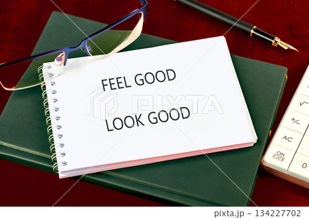 A spiral notepad shows the phrase FEEL GOOD LOOK GOOD. Nearby, glasses rest beside a black fountain pen, books, and a calculator on a dark burgundy surface A spiral notepad shows the phrase FEEL GOOD LOOK GOOD. Nearby, glasses rest beside a black fountain pen, books, and a calculator on a dark burgundy surface 134227702