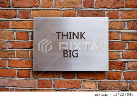 Words encouraging big dreams are engraved on a sleek metal sign, beautifully contrasting with the warm tones of a brick wall, inviting thoughts of innovation and growth 134227846