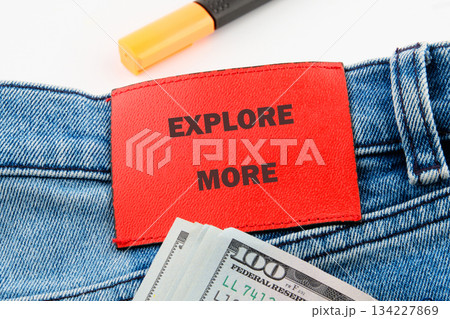 A vibrant red tag with EXPLORE MORE sits on denim fabric, alongside a stack of cash, inviting curiosity and adventure on a bright day filled with possibilities A vibrant red tag with EXPLORE MORE sits on denim fabric, alongside a stack of cash, inviting curiosity and adventure on a bright day filled with possibilities 134227869