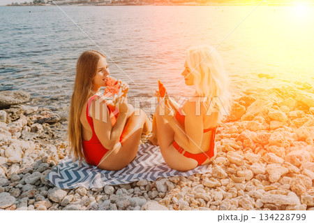 Watermelon Beach Summer: Two women enjoy refreshing watermelon slices on a rocky beach during summer vacation. 134228799