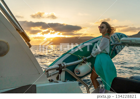 Woman, boat, ocean. Woman stands on a yacht with a paddleboard, enjoying a beautiful sunset. 134228821