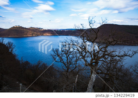 lake at the foot of a mountain surrounded by low mountains in autumn or winter surrounded by trees without leaves under a blue sky on the Edersee in Hesse Germany 134229419