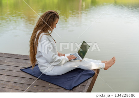 A woman is sitting on a dock with a laptop open in front of her. She is wearing white pants and a white sweater. The scene is peaceful and relaxing, with the water in the background. 134229580