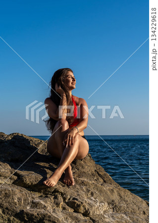 Woman relaxing on beach rocks, enjoying the sun by the sea under a clear blue sky with ample copy space Woman relaxing on beach rocks, enjoying the sun by the sea under a clear blue sky with ample copy space 134229618