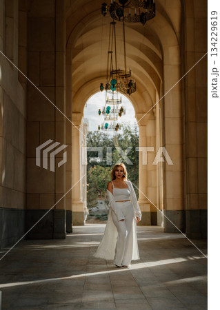 Woman, Architecture, Fashion - A woman in a white outfit walks through a grand arched hallway, showcasing stylish fashion in an architectural setting. 134229619