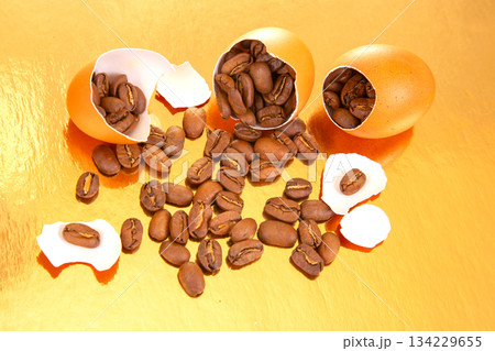 Creative still life of coffee beans emerging from a broken egg on a gold background 134229655