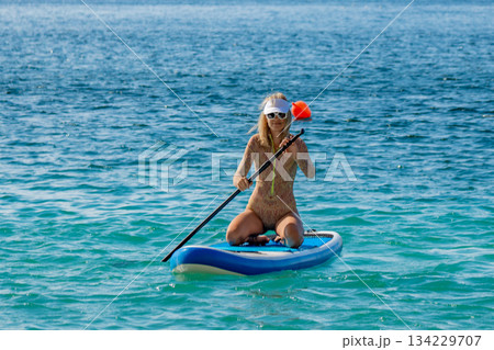 Paddleboarding, woman, ocean. Woman kneeling on stand-up paddleboard, paddling across blue ocean, enjoying summer recreation. 134229707