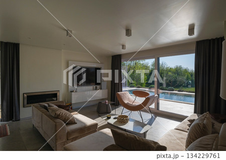 Modern luxury living room interior at sunset. Open-plan space featuring beige sofas, marble dining table, large glass windows with sun flare, and view of a patio. Stylish contemporary home design Modern luxury living room interior at sunset. Open-plan space featuring beige sofas, marble dining table, large glass windows with sun flare, and view of a patio. Stylish contemporary home design 134229761