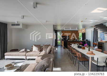 Modern luxury open-plan interior featuring a floating staircase, marble dining table, wooden kitchen, and beige sofa. Spacious contemporary home design with elegant lighting and gray tiled floors 134229763