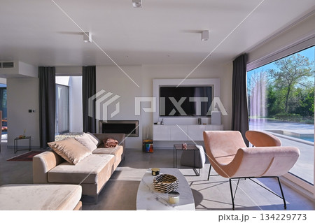 Modern luxury living room interior at sunset. Open-plan space featuring beige sofas, marble dining table, large glass windows with sun flare, and view of a patio. Stylish contemporary home design 134229773