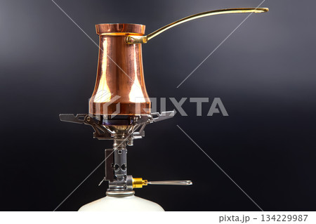 A shiny copper Turkish coffee pot with a brass handle sits atop a portable gas stove  134229987