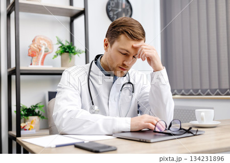 Handsome doctor man wearing medical uniform sitting on his workplace tired holding his head feeling fatigue and headache. Stress and frustration concept Handsome doctor man wearing medical uniform sitting on his workplace tired holding his head feeling fatigue and headache. Stress and frustration concept 134231896