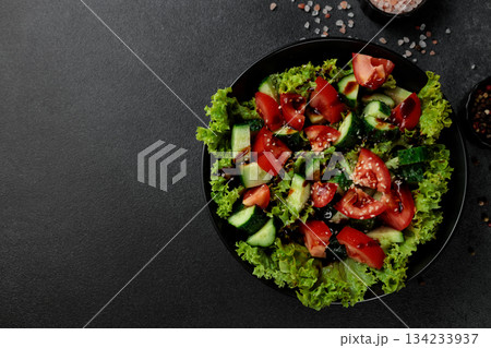 Vegetable salad of tomatoes, cucumbers, onions, lettuce dressed with aromatic dressing in a black plate on a gray background 134233937