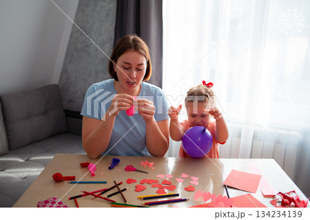 Mid shot of young Caucasian mother and her cute little daughter inflate balloons and make decorations for the holiday. Family sits at the table and makes festive decorations 134234139