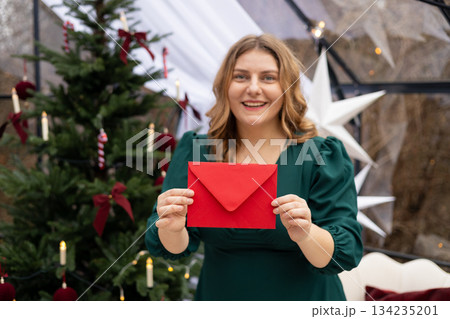 Young 30s woman getting Christmas greeting card from her beloved on background of xmas tree at cozy living room with festive interior. Surprised lady pleasure reading letter at home. 134235201