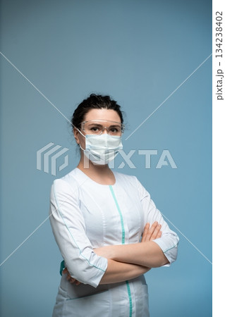 Confident female doctor in mask and protective glasses standing with folded arms on blue background Confident female doctor in mask and protective glasses standing with folded arms on blue background 134238402