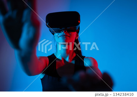 Woman extending arms forward toward virtual interface while wearing VR headset in neon sci-fi lighting 134238410