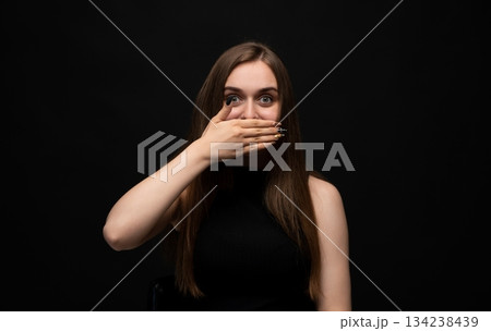 Surprised young woman covering mouth with hand in shocked emotional reaction on dark studio background Surprised young woman covering mouth with hand in shocked emotional reaction on dark studio background 134238439