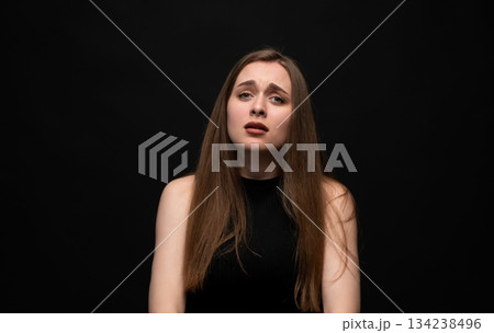 Sad young woman with worried facial expression in dramatic emotional studio portrait on dark background 134238496