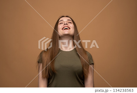 Joyful young woman laughing with head raised, expressive positive emotion portrait on warm studio background Joyful young woman laughing with head raised, expressive positive emotion portrait on warm studio background 134238563