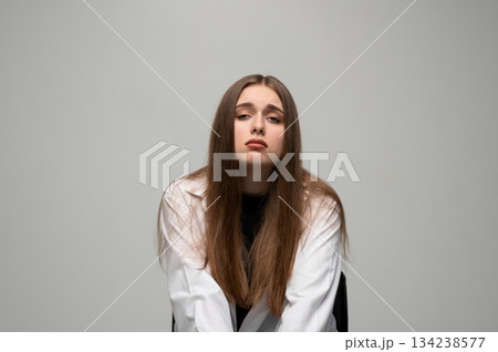 Sad tired woman in white coat looking down with bored facial expression on neutral gray studio background Sad tired woman in white coat looking down with bored facial expression on neutral gray studio background 134238577