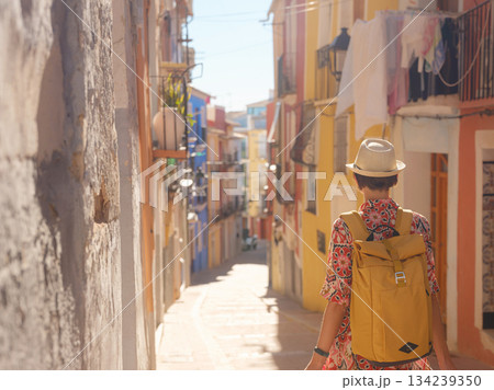 Woman in dress strolls through colorful streets of Spanish coastal town of La Vila Joiosa or Villajoyosa. sunny winter atmosphere highlights charm of Mediterranean architecture and quiet seaside life Woman in dress strolls through colorful streets of Spanish coastal town of La Vila Joiosa or Villajoyosa. sunny winter atmosphere highlights charm of Mediterranean architecture and quiet seaside life 134239350