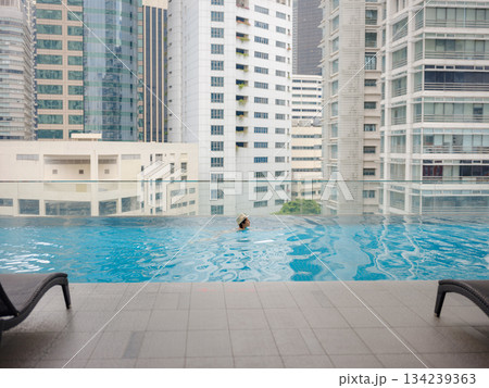 Young asian woman relaxing by pool at Kuala Lumpur hotel with view of surrounding skyscrapers, enjoying leisure time in vibrant urban setting. 134239363