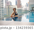Young asian woman relaxing by pool at Kuala Lumpur hotel with view of surrounding skyscrapers, enjoying leisure time in vibrant urban setting. 134239365