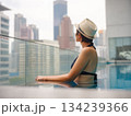 Young asian woman relaxing by pool at Kuala Lumpur hotel with view of surrounding skyscrapers, enjoying leisure time in vibrant urban setting. 134239366