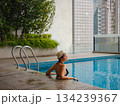 Young asian woman relaxing by pool at Kuala Lumpur hotel with view of surrounding skyscrapers, enjoying leisure time in vibrant urban setting. 134239367