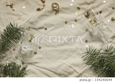 Golden star confetti tinsel, beige linen table cloth background. Champagne glasses. Fir branches floral frame, copy space. Christmas party concept. New Years flatlay, top view. Festive holiday banner 134240044