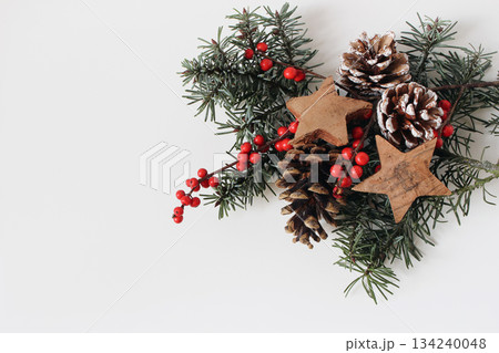 Christmas festive still life. Decorative composition. Pine cones, red holly berries, wooden stars, fir tree branches isolated on white table background. Winter floral flat lay, top view. Copy space Christmas festive still life. Decorative composition. Pine cones, red holly berries, wooden stars, fir tree branches isolated on white table background. Winter floral flat lay, top view. Copy space 134240048