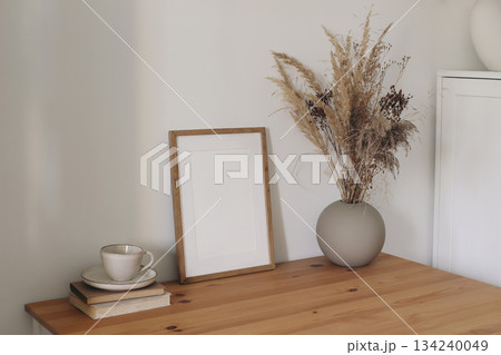 Minimal cozy interior scene with blank wooden picture frame mockup. Dried flowers, grass in vase. Coffee cup, books on table. Neutral Scandinavian home decor, natural rustic styling. Lifestyle design Minimal cozy interior scene with blank wooden picture frame mockup. Dried flowers, grass in vase. Coffee cup, books on table. Neutral Scandinavian home decor, natural rustic styling. Lifestyle design 134240049